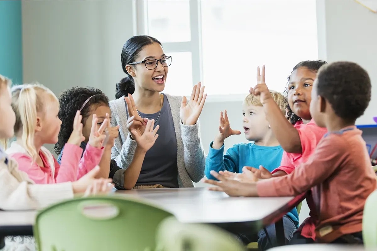 A teacher sits with her students at a table and leads them in a learning song.